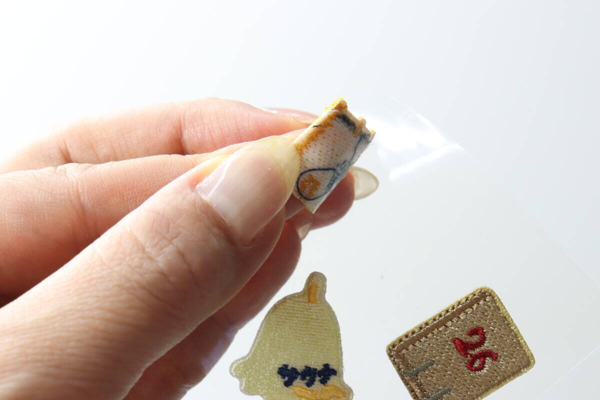 一隻手用手指從透明紙上撕下一塊桑拿主題的貼片。 指でサウナモチーフのワッペンを透明なシートから剥がしている手元