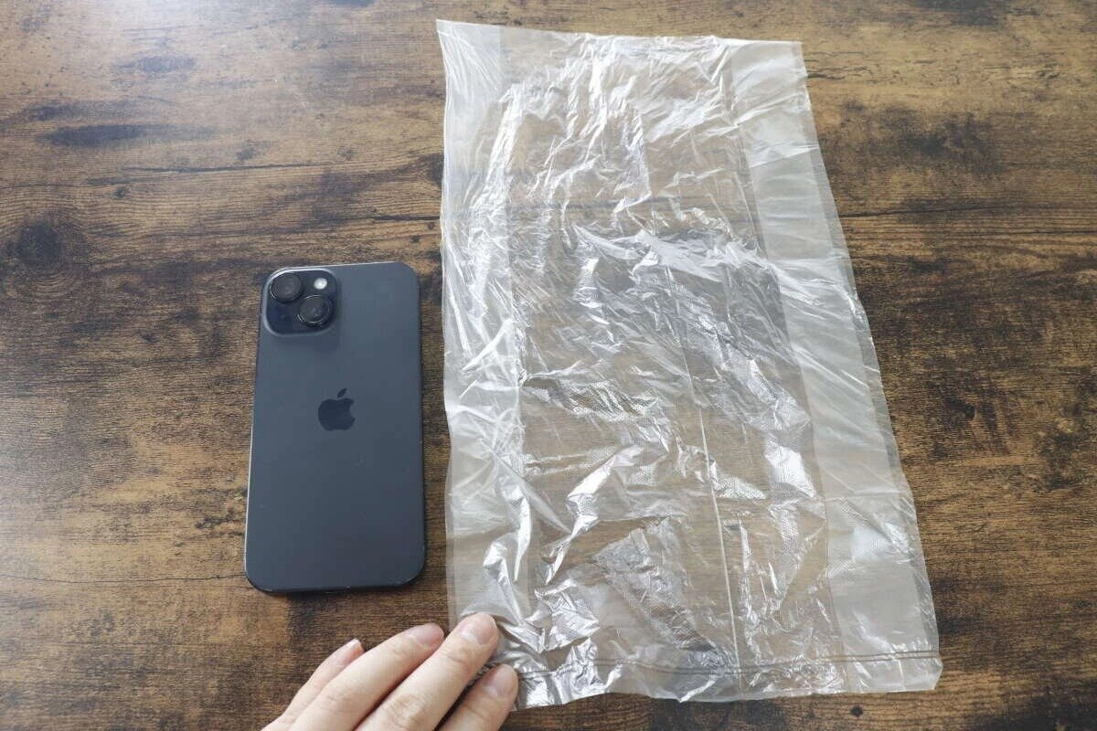 一部黑色智慧型手機放在木紋桌上,旁邊攤開著一套透明的廚房用品。 (尺寸對比) 木目調のテーブルに置かれた黒いスマートフォンと、その横に広げられた透明なキッチンパック。サイズ比較