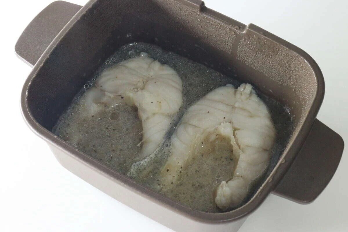 將兩塊浸沒在高湯中的魚片放入微波爐多功能烹飪鍋中。 レンジ調理マルチクッカーの中に煮汁に浸かった魚の切り身が2切れ
