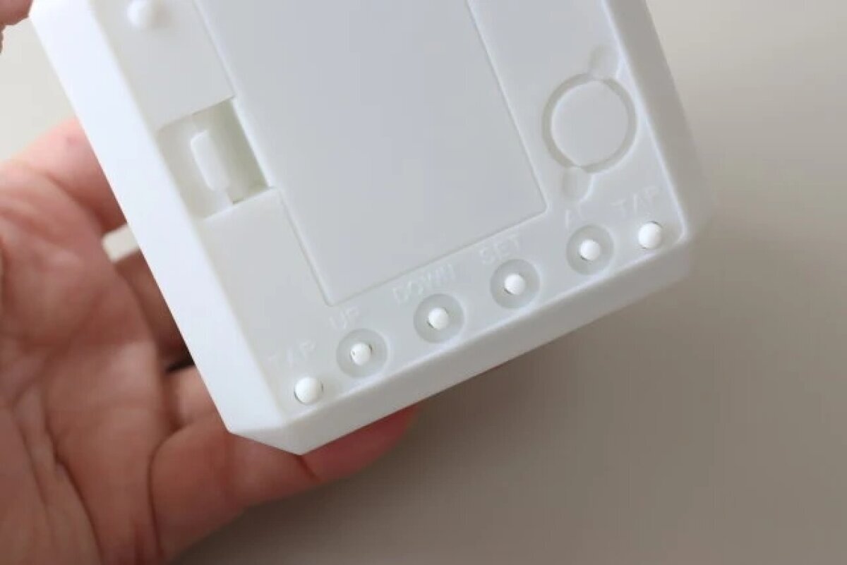 一個人用手指按壓白色「7色變色數字鐘」底部的設定鈕。 白い「7色変色ライト付デジタル時計」の底面にある設定ボタンを指で押している様子