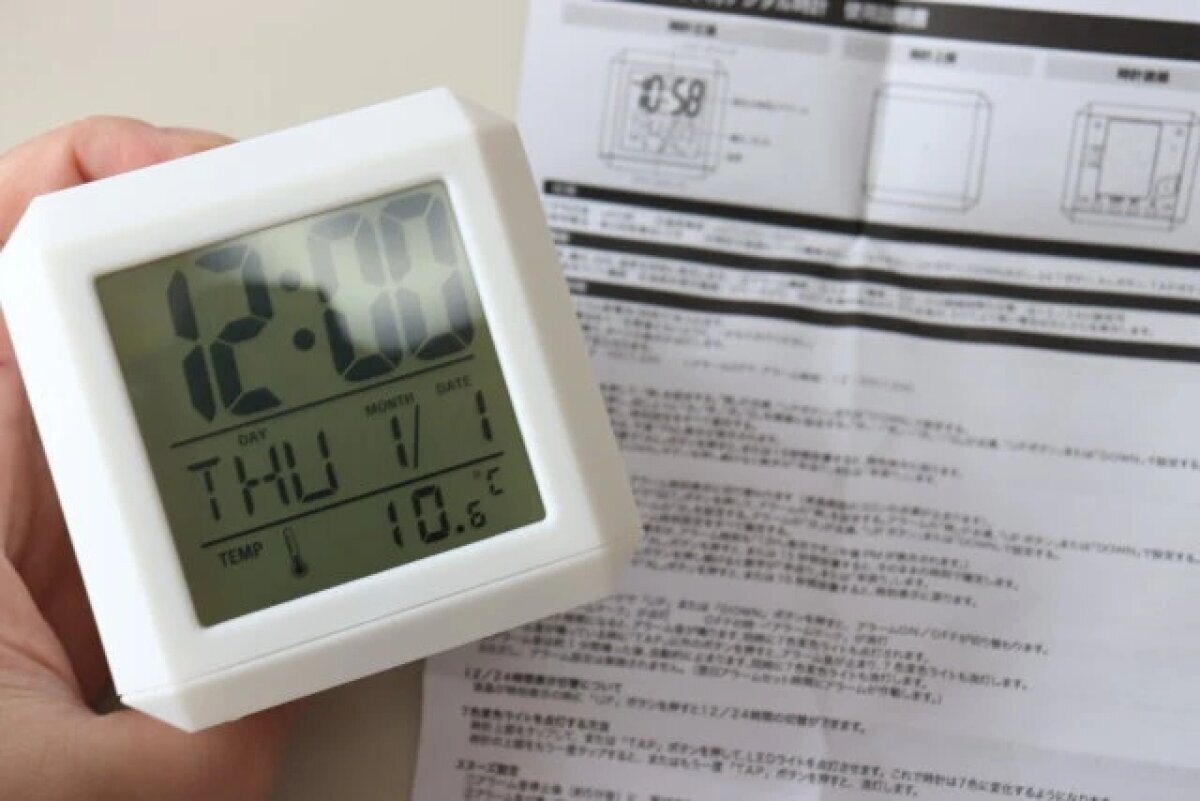 一手拿著一個白色的“帶有7種顏色變化燈光的數字時鐘”,另一手攤開著說明書。 白い「7色変色ライト付デジタル時計」を手に持ち、説明書を広げた状態