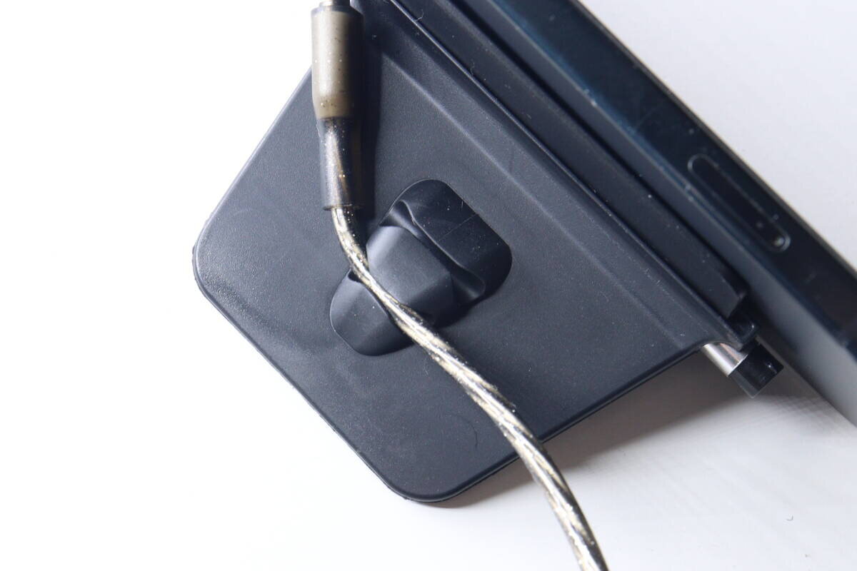 充電線夾在黑色智慧型手機支架背面的線夾上。 黒いスマホスタンドの背面にあるコードクリップに、充電ケーブルが挟まれている様子。