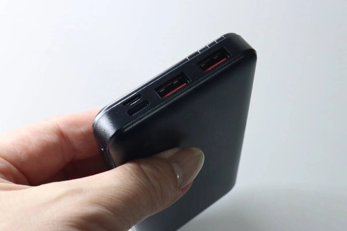 一個人一手拿著一塊黑色的行動電源,側面有多個充電接口。 黒いモバイルバッテリーを片手で持っている様子。側面には複数の充電ポートがある