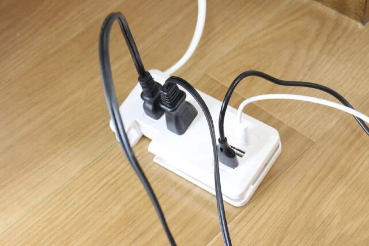 木地板上放著一個白色電源插座,上面插著幾根黑色電線。 白いコンセントタップがフローリングの床に置かれ、複数の黒いケーブルが差し込まれている