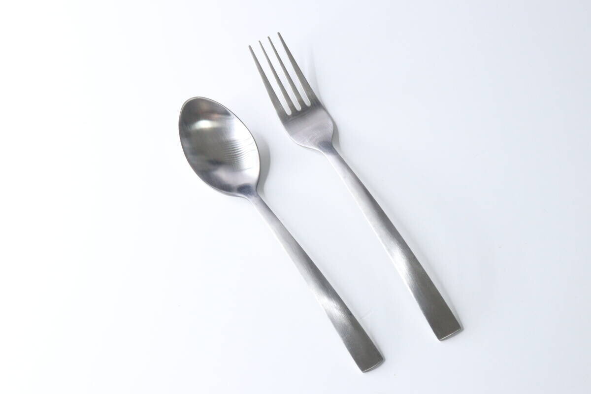 一把簡單的不銹鋼甜點勺和一把甜點叉斜放在白色背景上。 白い背景に、シンプルデザインのステンレス製デザートスプーンとデザートフォークが斜めに置かれている