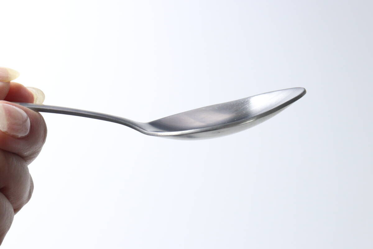 照片顯示的是手指握著的不銹鋼甜點勺側面的特寫,露出了湯匙尖的弧度。 指で持ったステンレス製デザートスプーンの側面がアップで写っており、先端のカーブがわかる