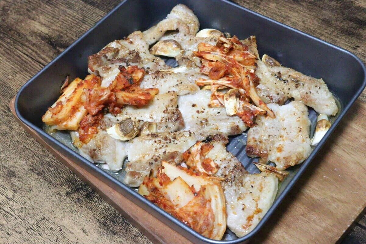 這道菜的特色在於將豬肉放在烤盤上烤製,上面放上泡菜和大蒜。 オーブントレーで焼かれた豚肉とキムチ、にんにくが盛り付けられている