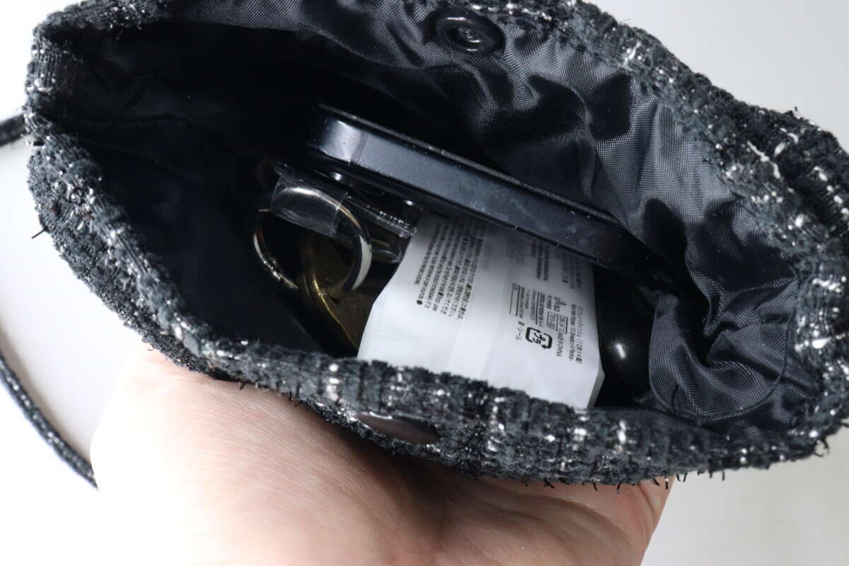 從上方俯視,可以看到黑色粗花呢單肩包裡裝著智慧型手機、鑰匙和其他物品。 黒いツイードショルダーバッグの中にスマホや鍵などの荷物が入っている様子を上から覗き込んでいる