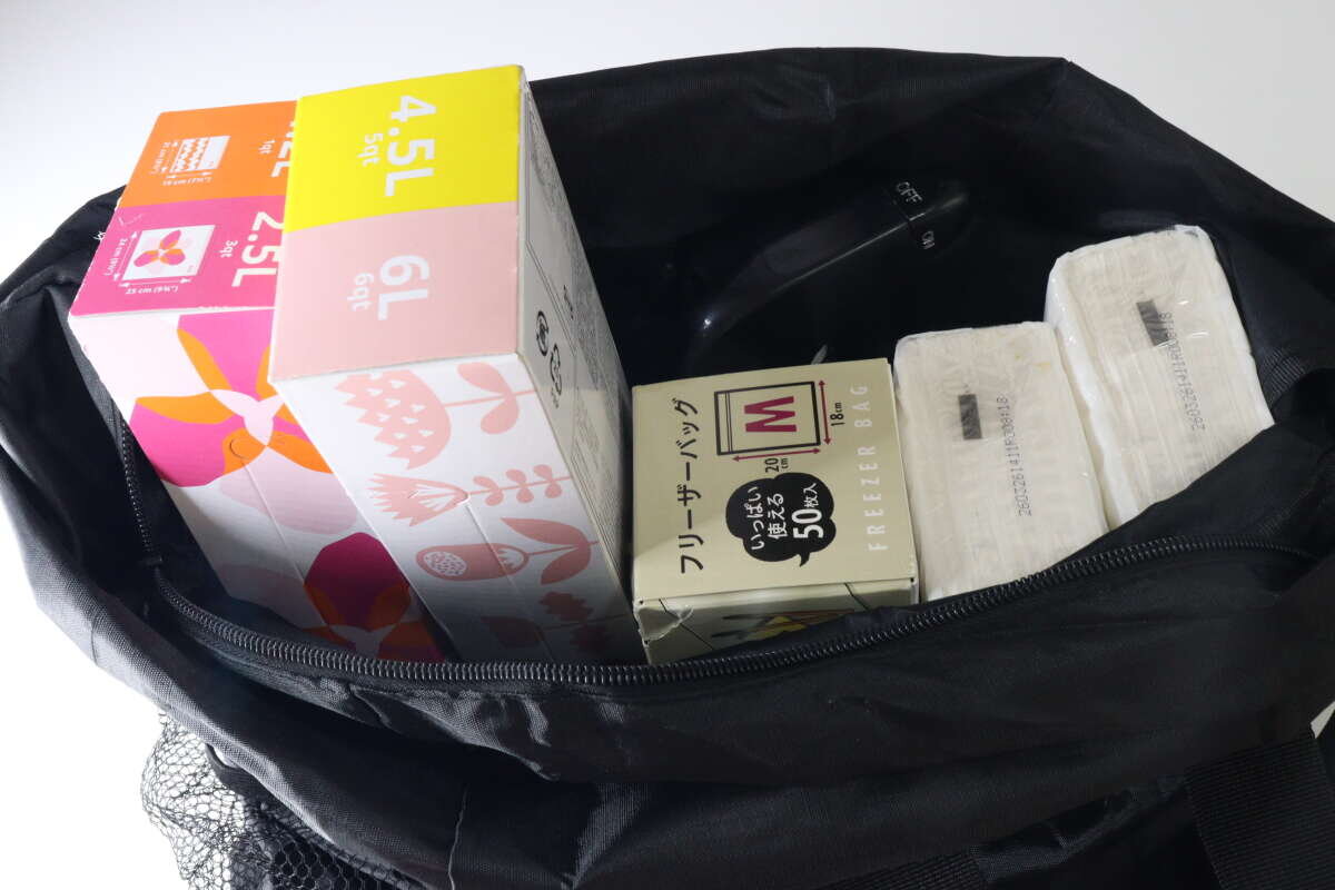 在展開的黑色手提背包裡,可以看到一包軟紙巾、一盒拉鍊袋和一瓶除臭噴霧。 広げられた黒いトート型バックパックの中に、ソフトパックティッシュ、箱入りジップ付きバッグ、消臭スプレーが収納されている様子。