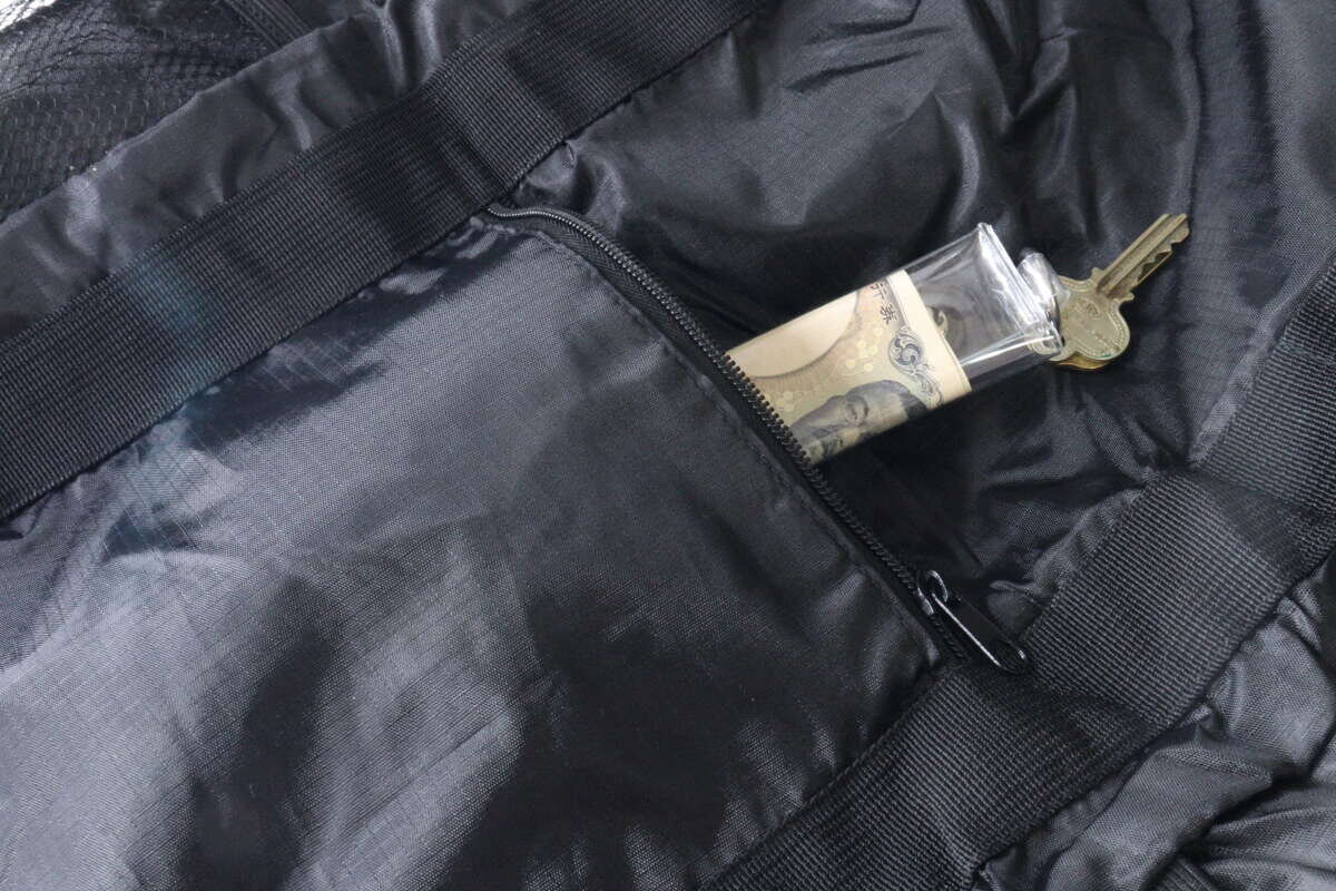 黑色手提式背包正面拉鍊口袋裡存放著一個裝有鈔票和鑰匙的透明盒子。 黒いトート型バックパックの前面にあるファスナーポケットに、お札と鍵が入った透明なケースが収納されている。