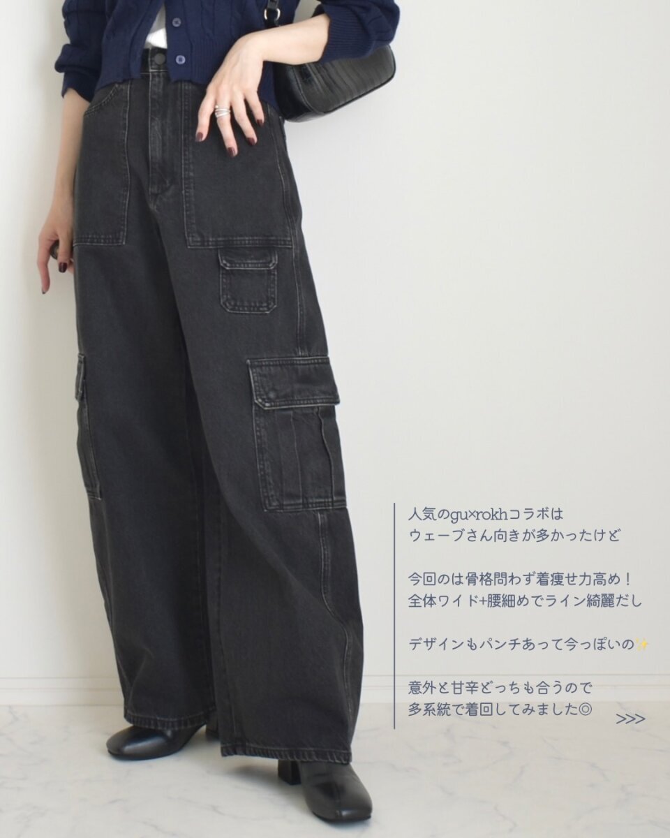 一位身穿黑色工裝牛仔褲和海軍藍開襟衫的女子全身照,她手提一個黑色包包。 黒いバレルユーティリティジーンズとネイビーのカーディガンを着用し、黒いバッグを持つ女性の全身