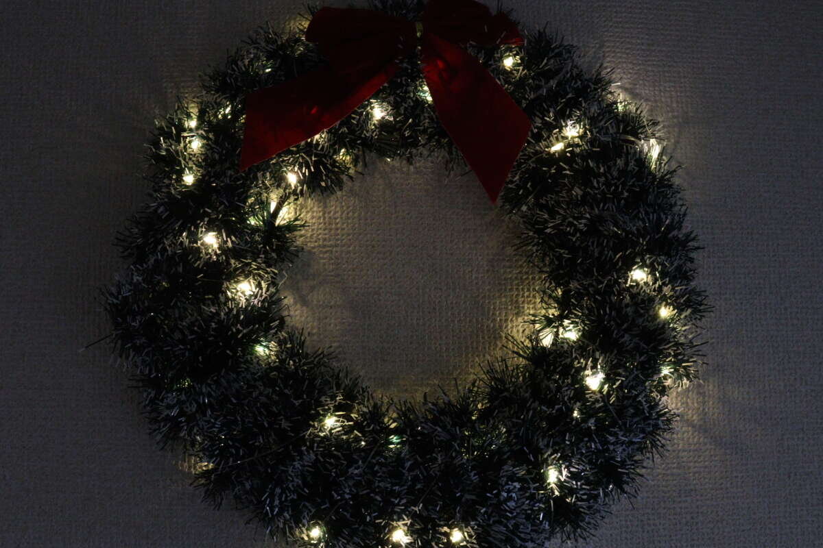 一個纏繞著彩燈的聖誕花環,在昏暗的房間裡被點亮。 クリスマスリースにストリングライトを巻き付け、暗い部屋で点灯させた様子。
