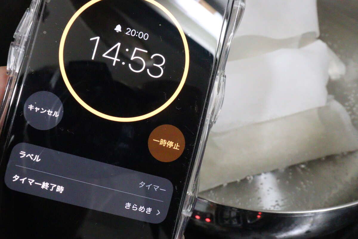 圖片顯示有人將智慧型手機的計時器螢幕放在鍋上方,以測量水沸騰的時間。 茹で時間を測るためにスマートフォンのタイマー画面を鍋にかざしている様子
