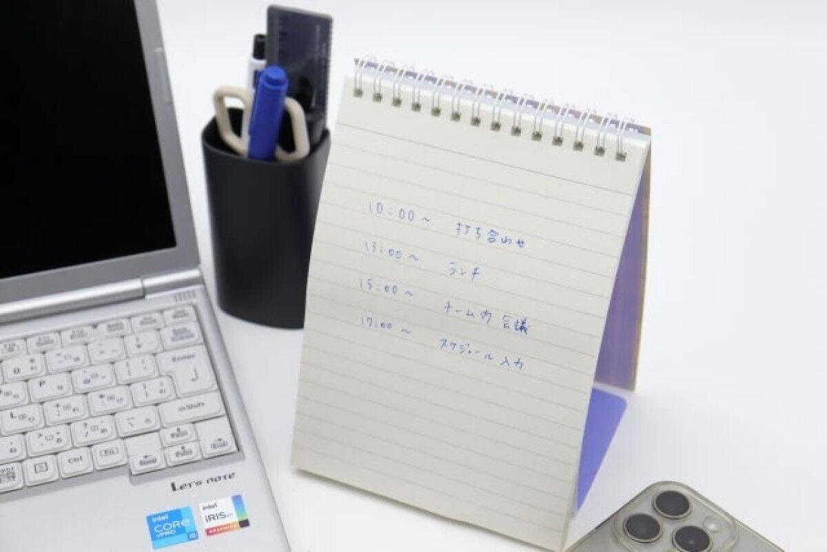 電腦旁邊支著一個直立式筆記本,裡面寫著行程。 PCの横にスケジュールが書き込まれたスタンドリングノートが立てられている様子