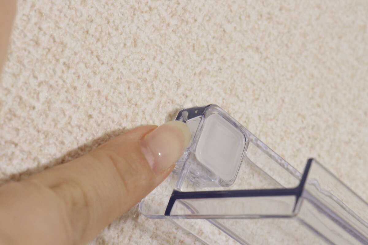 這個人正用手指牢牢抓住安裝在牆上的透明架子的四個角,以將其固定到位。 壁に設置した透明な棚の角を指でしっかりと押さえて固定している様子