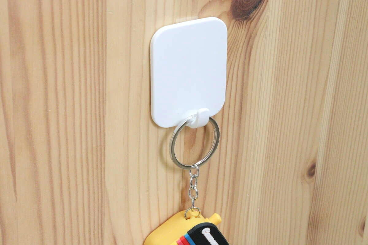 木目調の壁に透明な伸びるテープで固定された白いフックに、黄色いキーホルダー付きの鍵が吊り下げられている 木目調の壁に透明な伸びるテープで固定された白いフックに、黄色いキーホルダー付きの鍵が吊り下げられている
