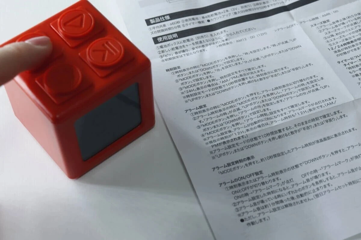 赤いブロック型デジタルタイマー時計のボタンを指で押しながら説明書を見ている 赤いブロック型デジタルタイマー時計のボタンを指で押しながら説明書を見ている