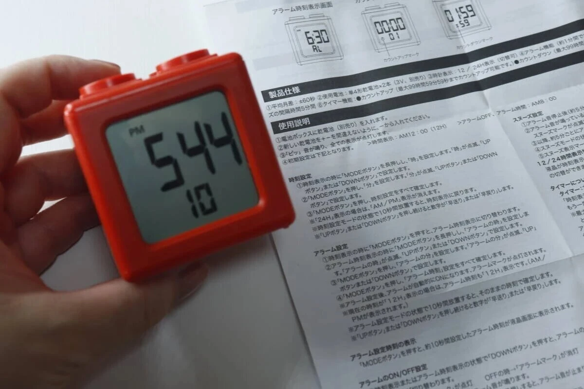 赤いブロック型デジタルタイマー時計の画面に「5:44 PM」と表示され、説明書が横にある 赤いブロック型デジタルタイマー時計の画面に「5:44 PM」と表示され、説明書が横にある