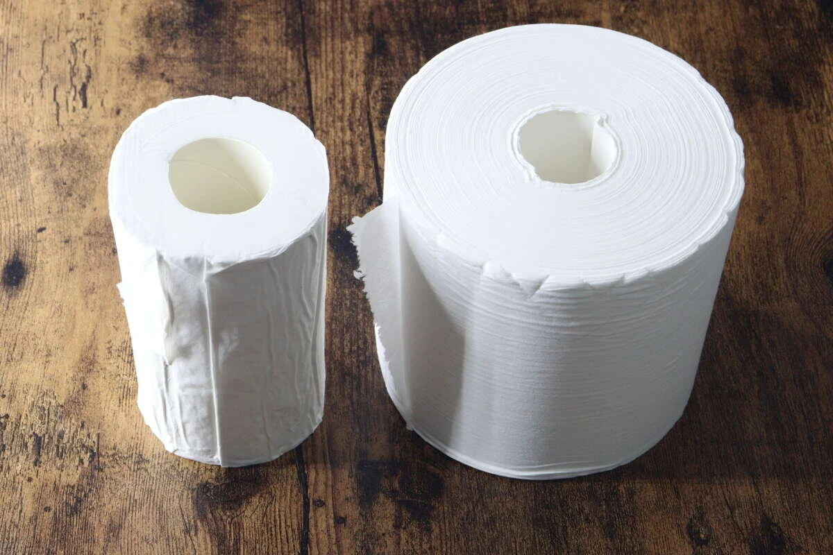 木目のテーブルに、左に圧縮トイレットペーパー、右に通常の5倍巻きトイレットペーパーが並べられている。 木目のテーブルに、左に圧縮トイレットペーパー、右に通常の5倍巻きトイレットペーパーが並べられている。