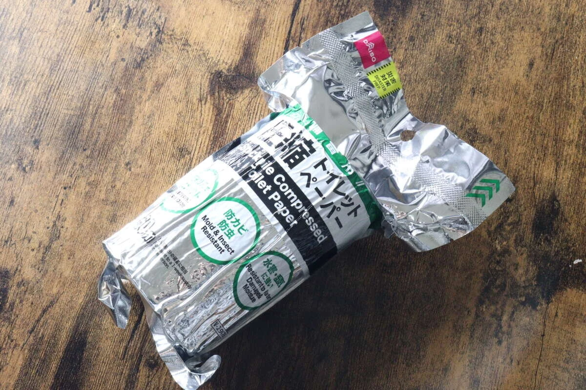 開封途中のアルミ包装に入ったダイソーの備蓄用圧縮トイレットペーパーが木目のテーブルに置かれている。 開封途中のアルミ包装に入ったダイソーの備蓄用圧縮トイレットペーパーが木目のテーブルに置かれている。