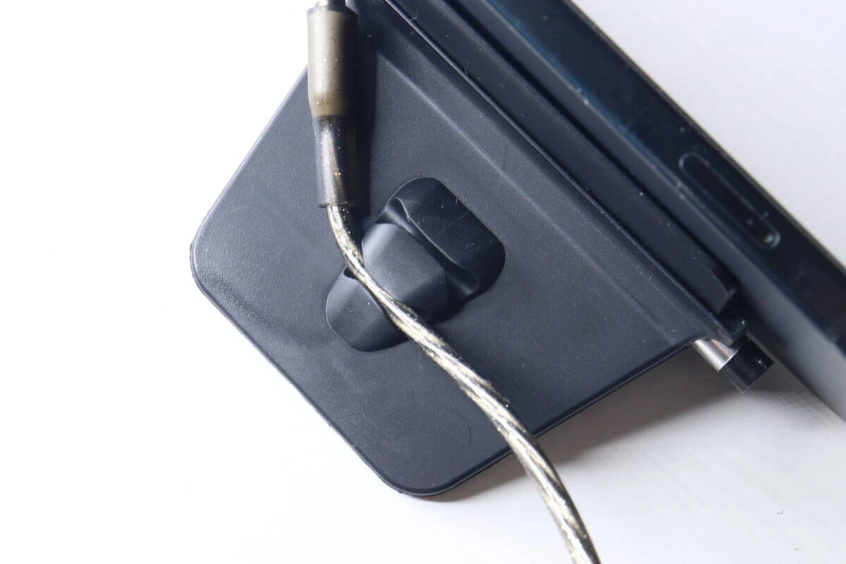 黒いスマホスタンドの背面にあるコードクリップに、充電ケーブルが挟まれている様子。 黒いスマホスタンドの背面にあるコードクリップに、充電ケーブルが挟まれている様子。