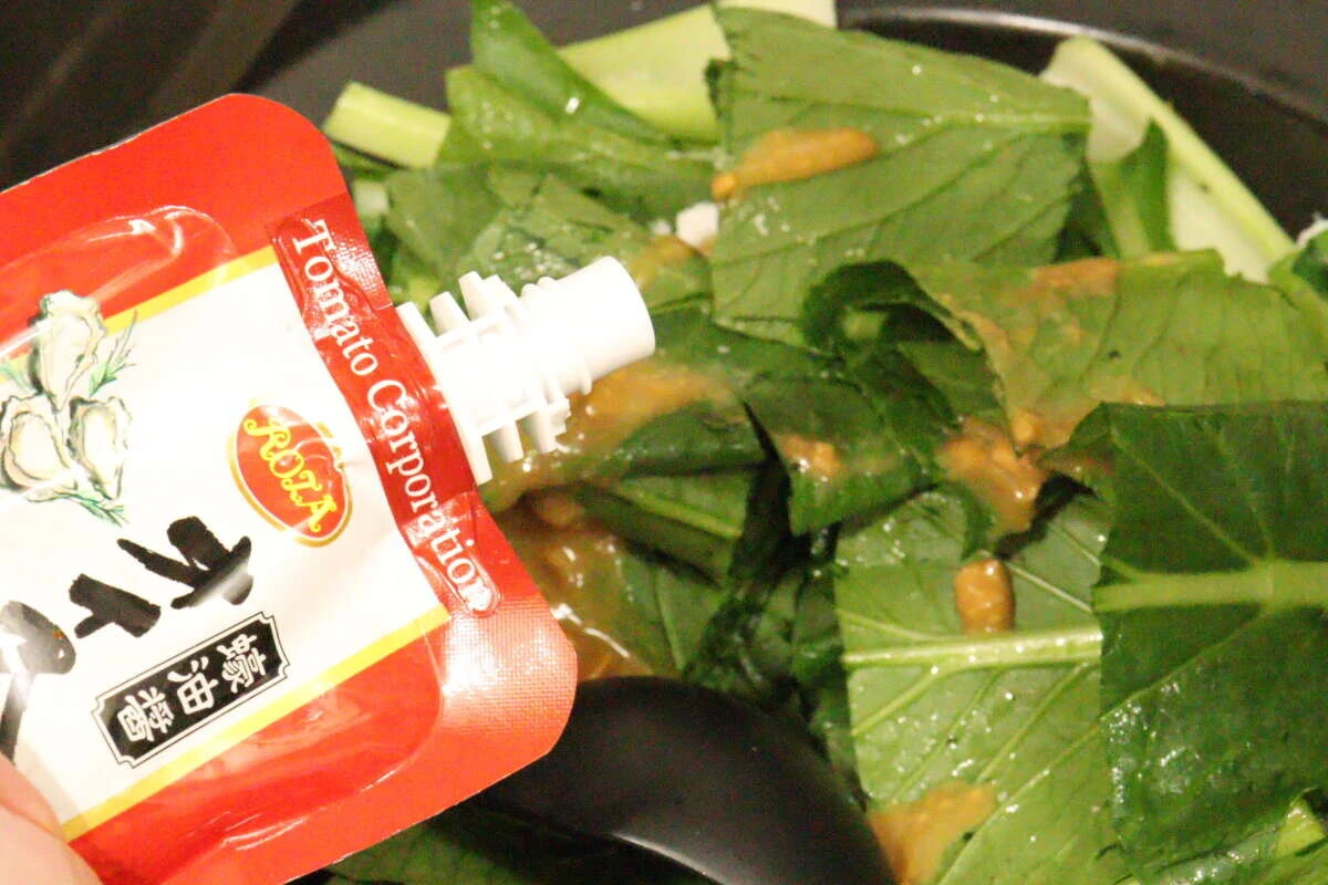 フライパンで炒めている青菜に、ダイソーのオイスターソースのパウチからソースを注ぎ入れている手元 フライパンで炒めている青菜に、ダイソーのオイスターソースのパウチからソースを注ぎ入れている手元