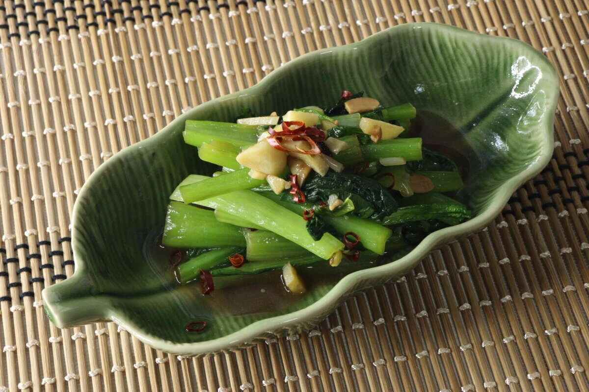 緑色の葉っぱ型のお皿に盛り付けられた、青菜とニンニク、唐辛子の炒め物が竹製のランチョンマットの上に置かれている 緑色の葉っぱ型のお皿に盛り付けられた、青菜とニンニク、唐辛子の炒め物が竹製のランチョンマットの上に置かれている