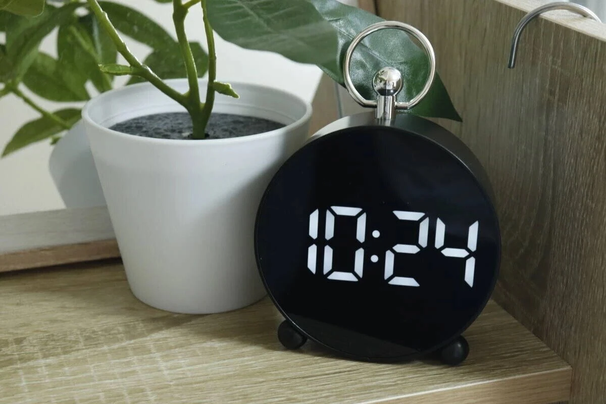 黒い丸型LEDデジタル時計が木製棚に置かれ、隣に白い鉢植えの観葉植物がある 黒い丸型LEDデジタル時計が木製棚に置かれ、隣に白い鉢植えの観葉植物がある
