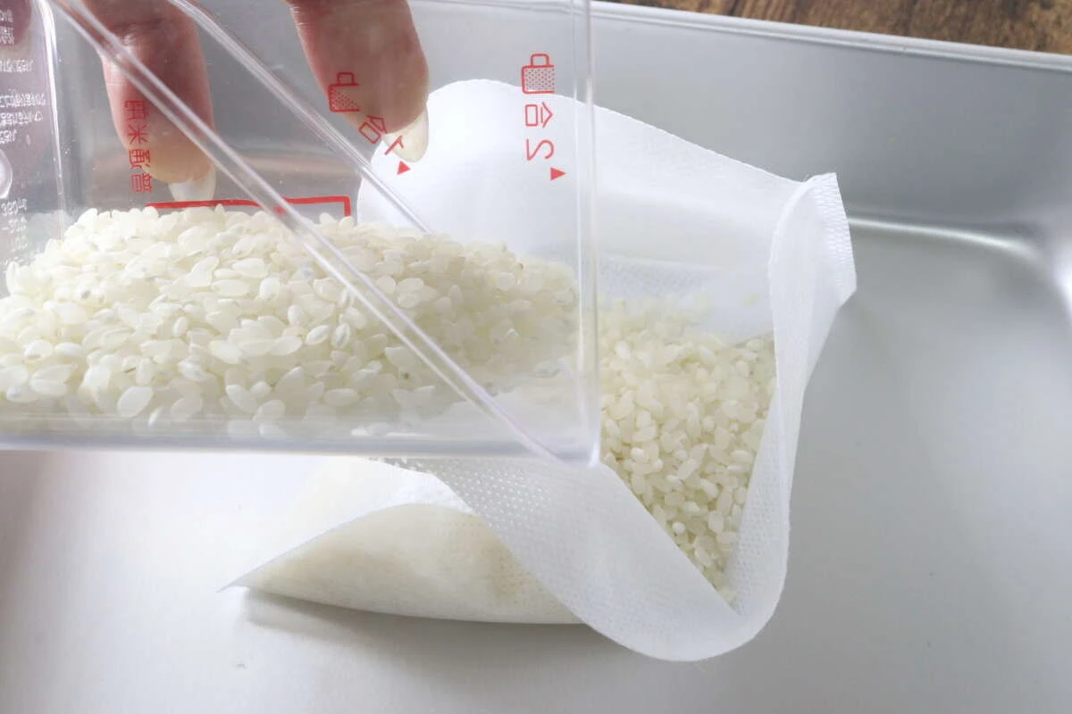 計量カップから白い生米を不織布の炊飯袋へ入れている工程 計量カップから白い生米を不織布の炊飯袋へ入れている工程