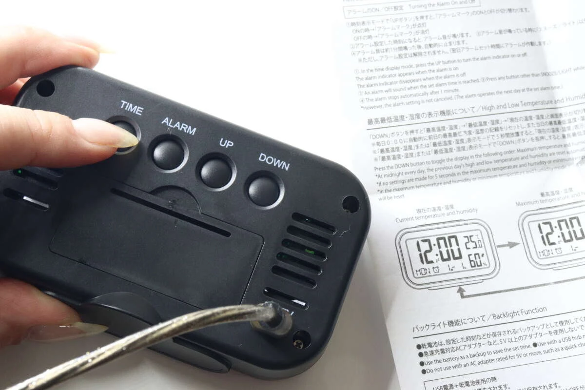 時計の裏側にあるボタンとUSB給電ケーブルの接続部分を手で支えている様子 時計の裏側にあるボタンとUSB給電ケーブルの接続部分を手で支えている様子