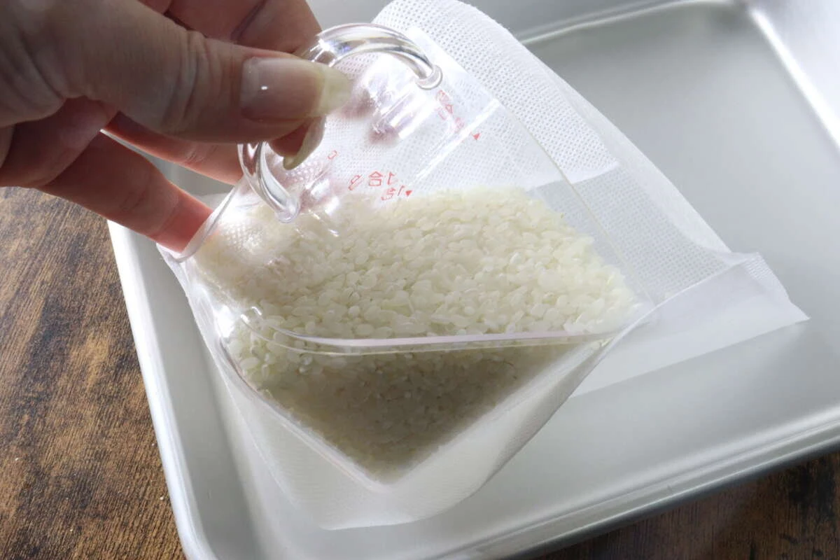 計量カップを使って生米を白い不織布製の炊飯袋に入れている様子 計量カップを使って生米を白い不織布製の炊飯袋に入れている様子