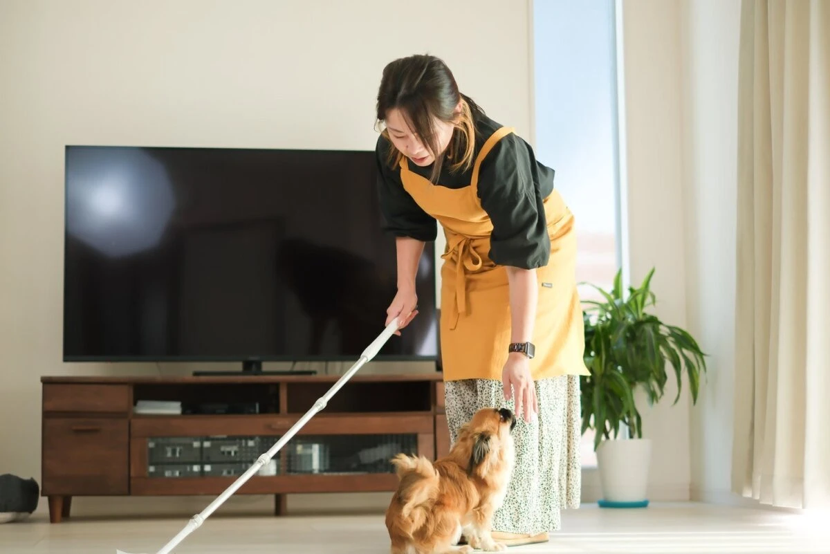 黄色いエプロンを着た女性がフローリングワイパーを持ちながら犬と触れ合っている