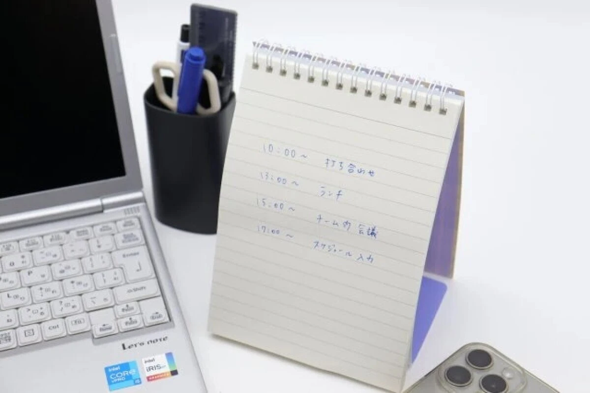 PCの横にスケジュールが書き込まれたスタンドリングノートが立てられている様子