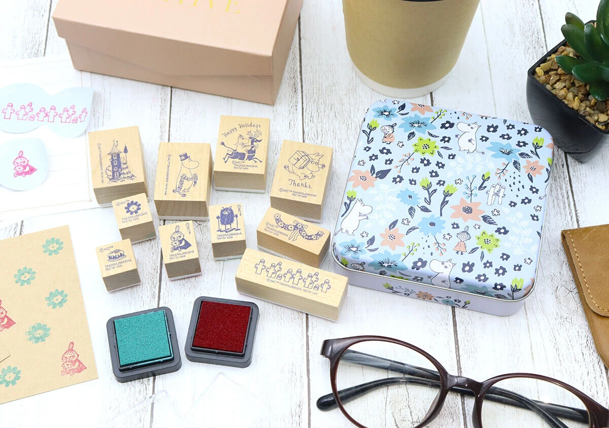 ムーミンの木製スタンプ10個と2色のインクパッド、花柄の缶ケース、眼鏡などが木製のテーブルに置かれている俯瞰。 ムーミンの木製スタンプ10個と2色のインクパッド、花柄の缶ケース、眼鏡などが木製のテーブルに置かれている俯瞰。