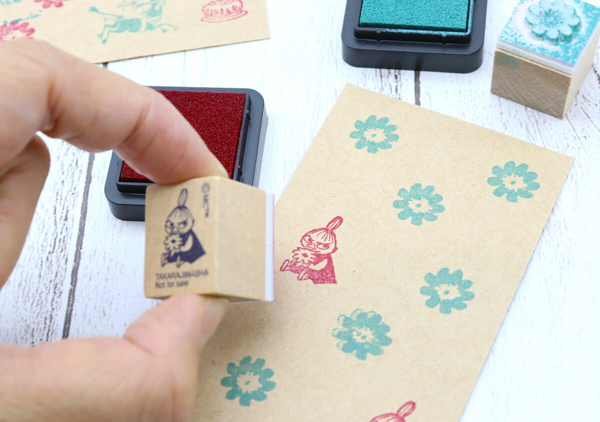 手がリトルミイのスタンプを持ち、クラフト紙に赤いインクで押している様子。紙には緑色の花柄スタンプも押されている。 手がリトルミイのスタンプを持ち、クラフト紙に赤いインクで押している様子。紙には緑色の花柄スタンプも押されている。
