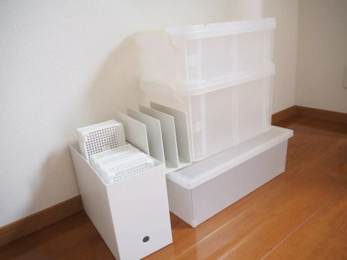 白い壁とフローリングの床に、様々なサイズの白い収納ケースやファイルボックスが積まれている