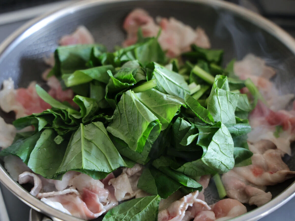 炒めている豚バラ肉の上にざく切りにした小松菜を入れている様子