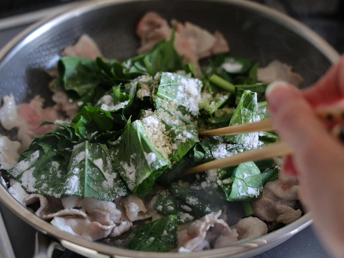 フライパンの小松菜と豚肉に砂糖と片栗粉を振りかけているところ