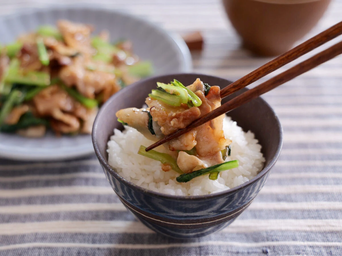 茶碗のご飯に箸で炒め肉を乗せている手元と背景の皿に盛られた料理